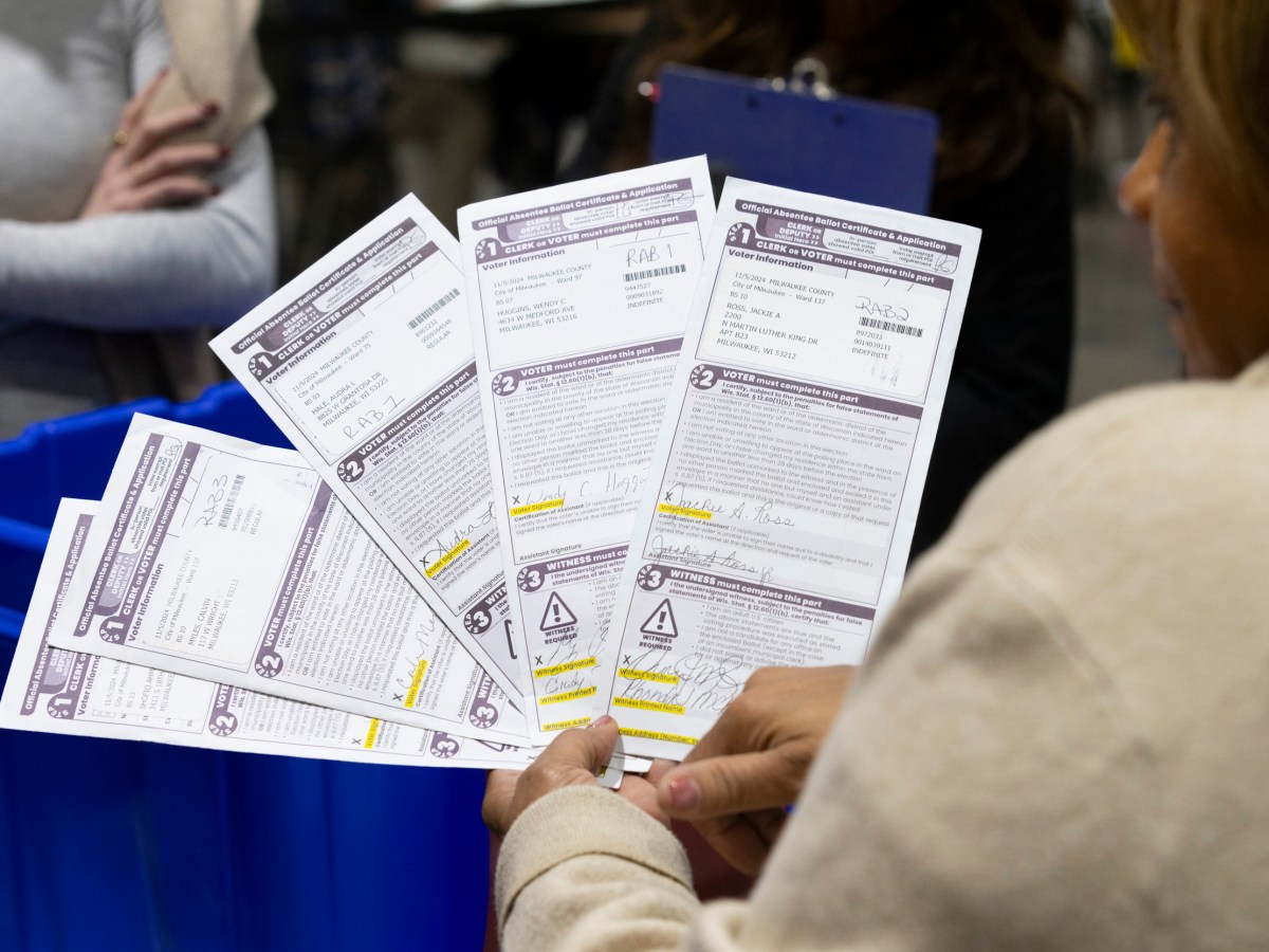 A person holds five absentee ballot forms near blue bins while others stand nearby.