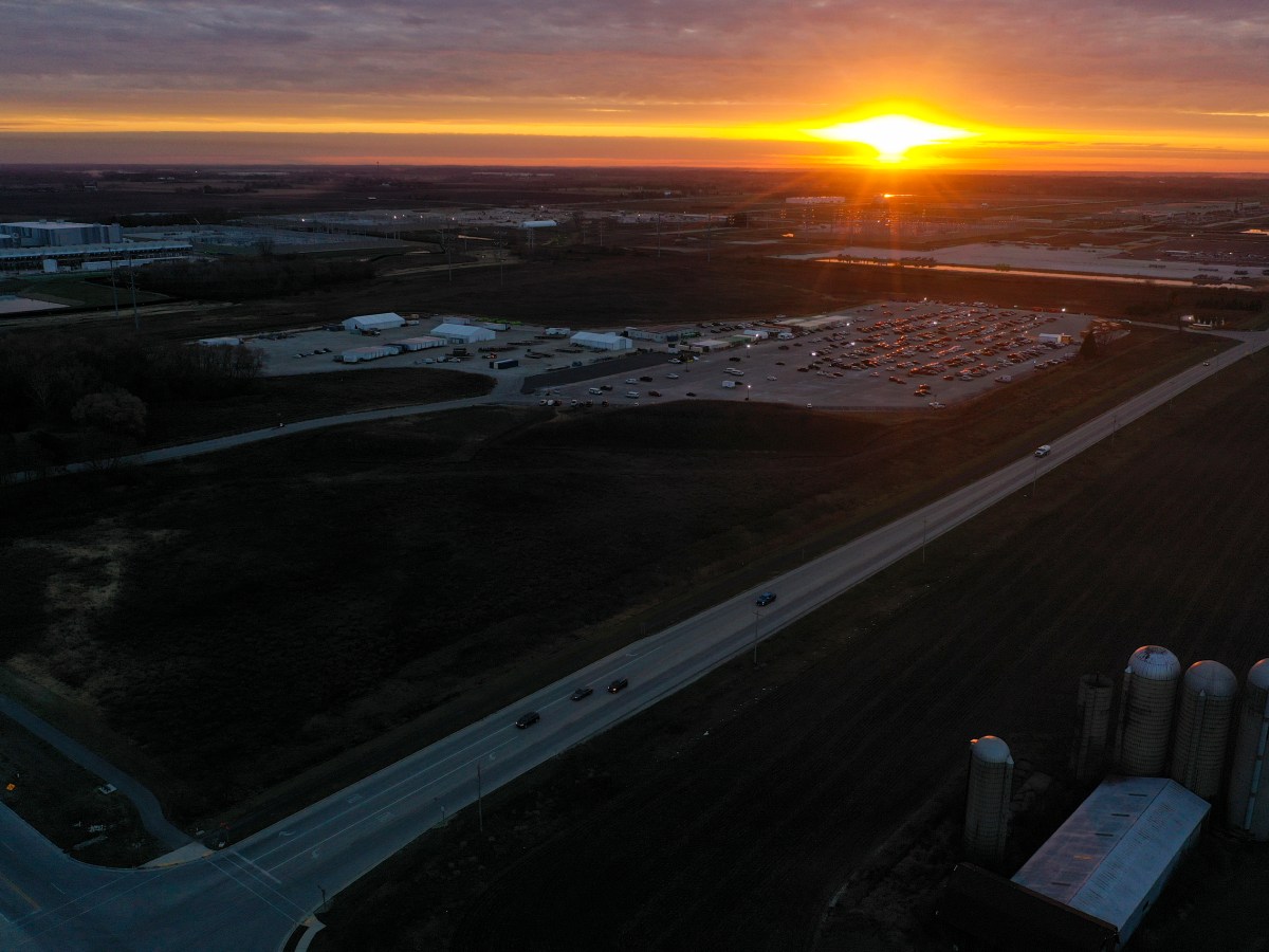 An aerial view of a large industrial complex next to a pond and surrounding construction areas at sunset, with orange light along the horizon under a cloudy sky.