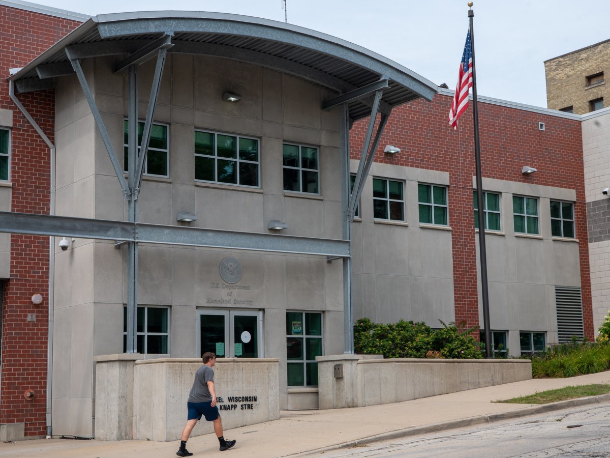 A person in shorts walks past a building labeled "U.S. Department of Homeland Security" with an American flag on a pole outside.
