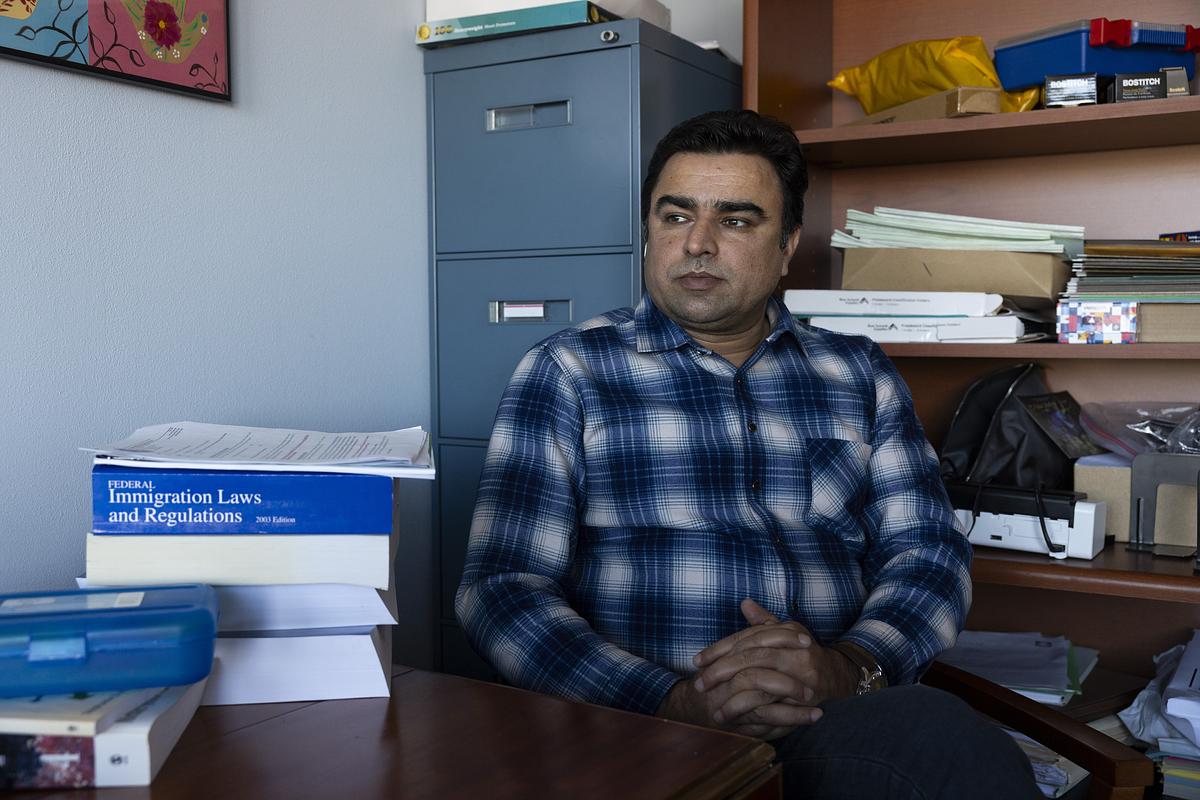 A person sits at a desk in an office, wearing a plaid shirt, with stacks of papers and books including one titled “Federal Immigration Laws and Regulations” nearby.