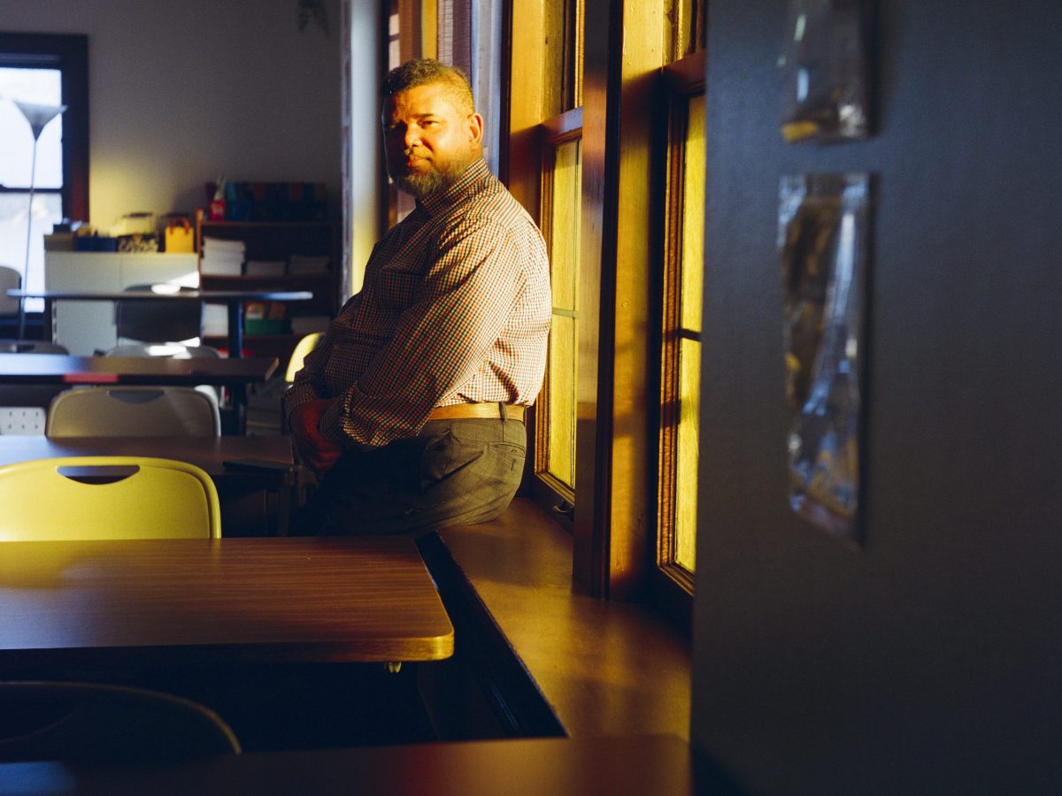 A person leans against a windowsill in a room, with sunlight streaming through tall windows and tables and chairs in the room.