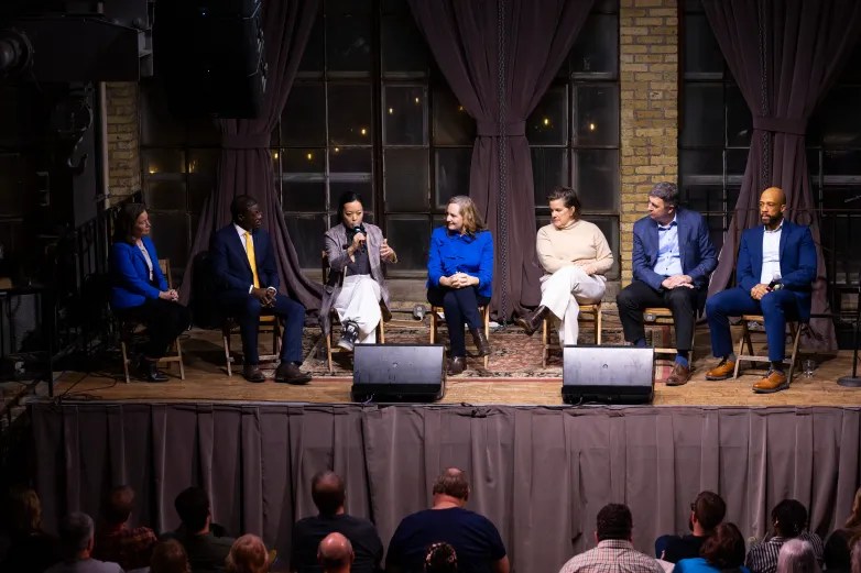 Seven people sit in a row on a stage as one speaks into a microphone, with an audience seated in front and large windows with curtains behind those on the stage.