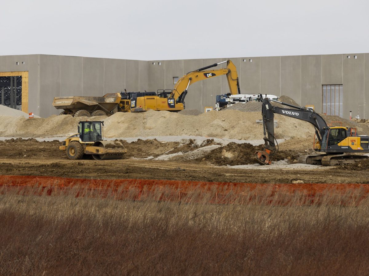 Construction equipment including excavators and a roller are on a dirt site with large piles of gravel in front of a concrete building.