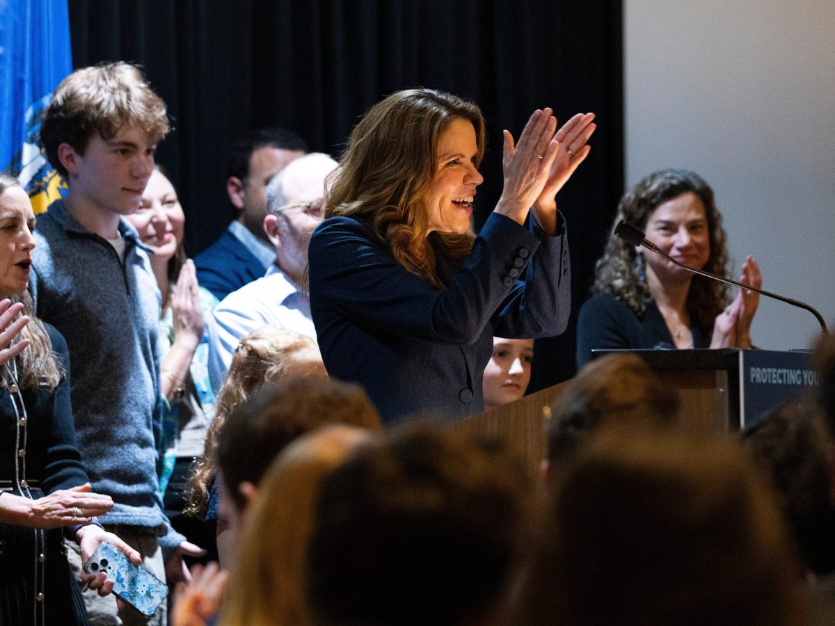 A person stands at a podium clapping while people behind the person and in an audience applaud in a crowded room.