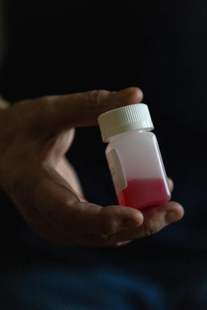 A person's hand holds a small bottle containing a red liquid against a dark background.
