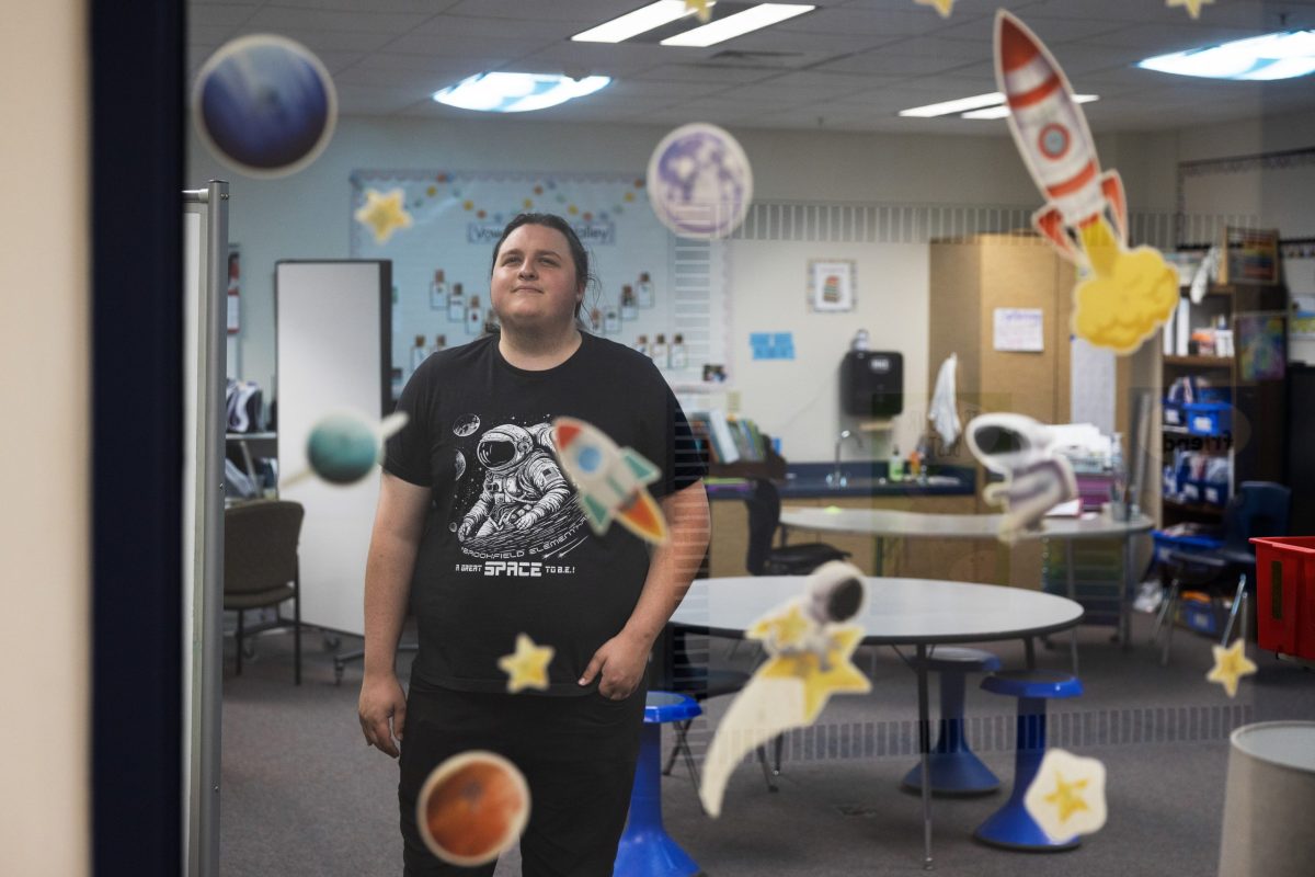 A person wearing a T-shirt with an astronaut graphic stands in a classroom decorated with paper planets, stars and rockets on a glass wall.