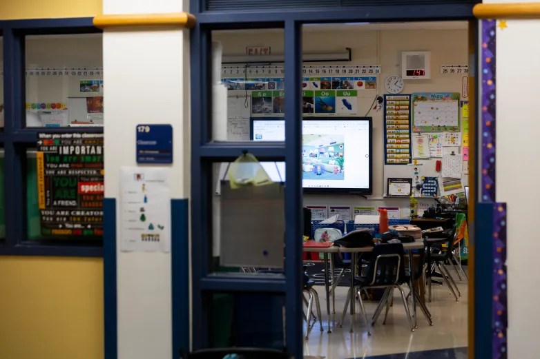 An empty classroom with desks, posters and a wall-mounted screen is visible through windows and an open doorway with a sign marked "179" on the wall outside the room.