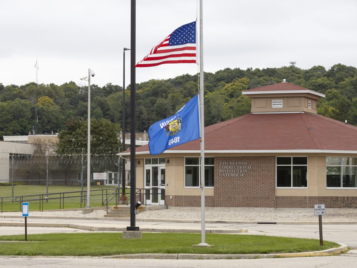 An American flag and a Wisconsin flag are attached to a pole outside a building labeled “Taycheedah Correctional Institution Gatehouse,” with fencing and trees in the background.