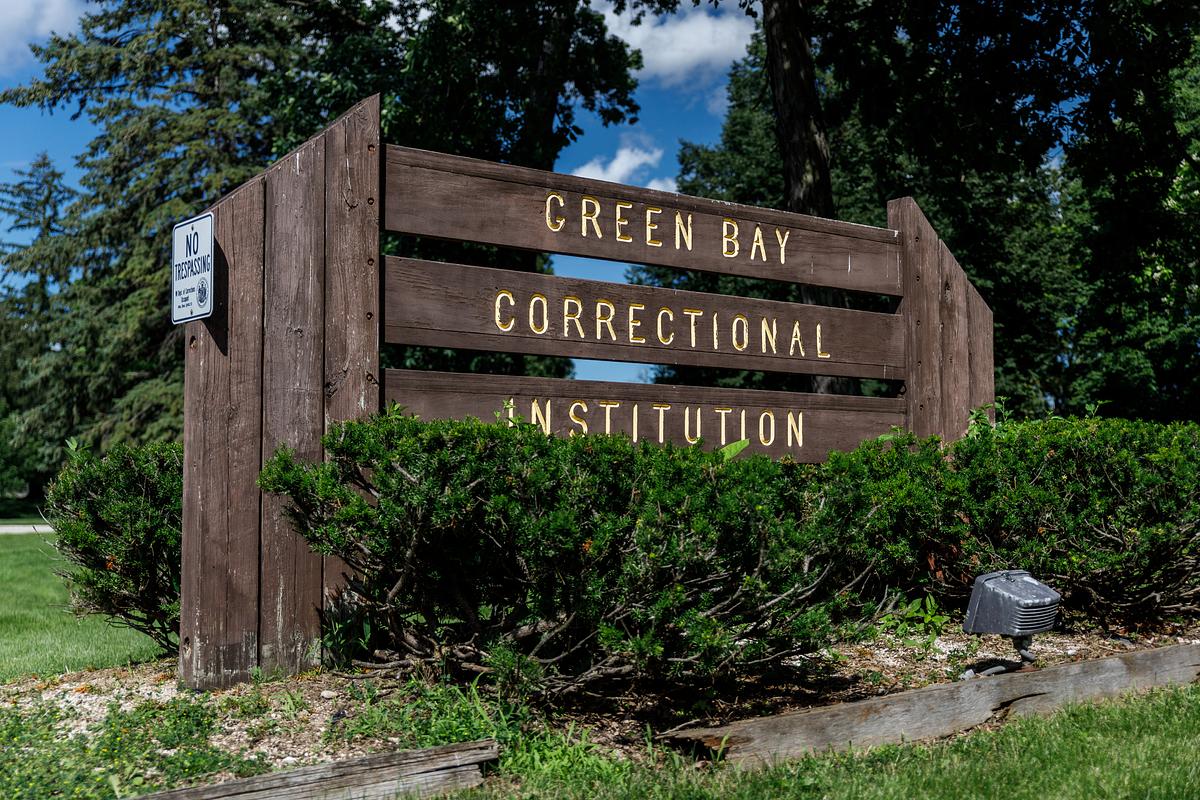 Wooden sign with yellow lettering reads "Green Bay Correctional Institution" beside a smaller "No trespassing" sign, surrounded by green shrubs and trees under a blue sky.