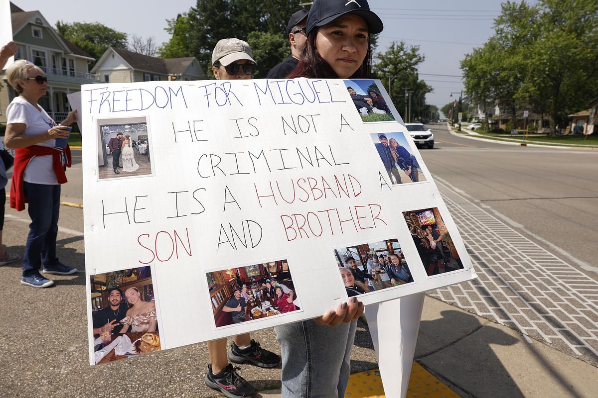 A person holds a sign reading "Freedom for Miguel he is not a criminal he is a husband son and brother" with photos attached, while others stand along a street nearby.