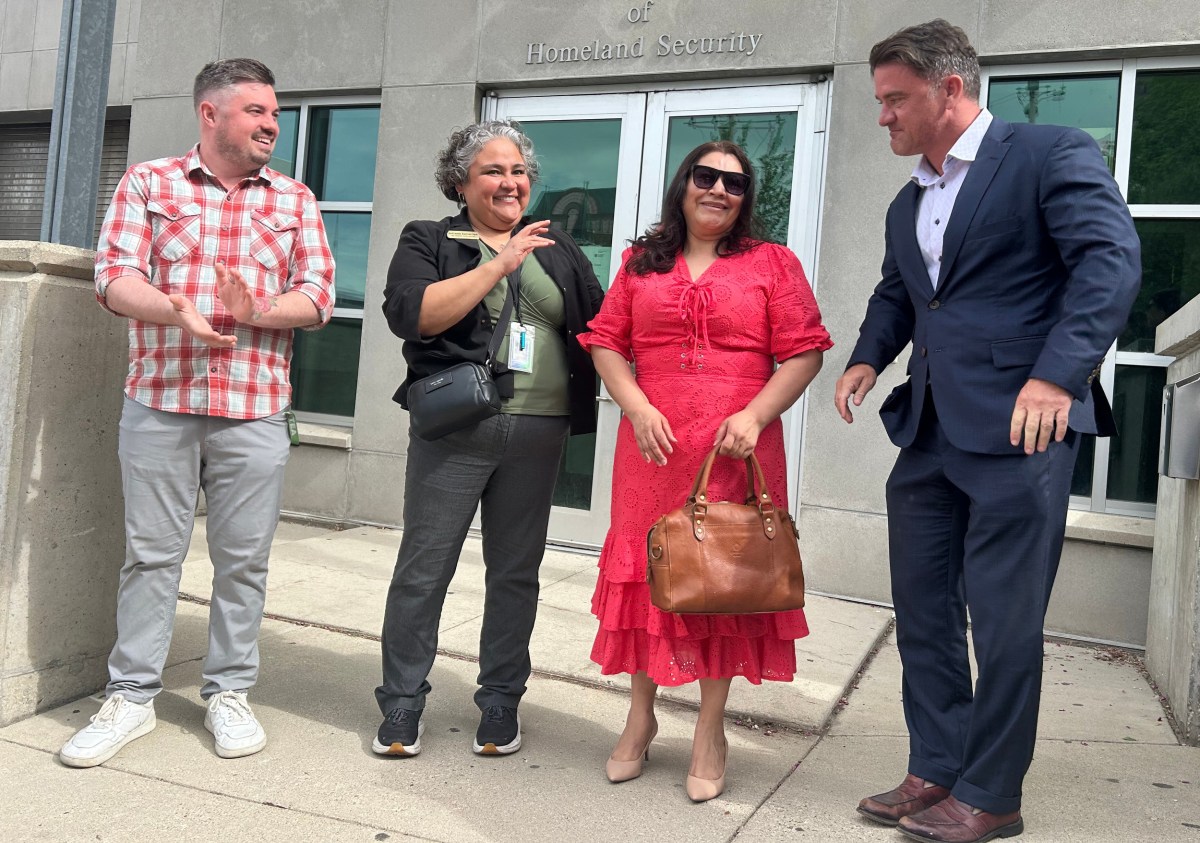 Four people stand on a sidewalk outside a building entrance with signage reading "Homeland Security." One person wearing a red dress holds a brown handbag.