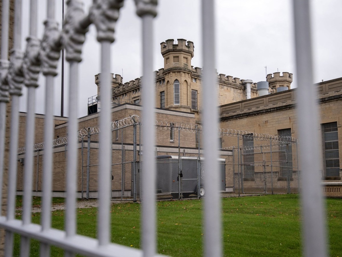 Metal fence in foreground with view of a tan brick building topped with guard towers, barred windows and fencing lined with razor wire under an overcast sky